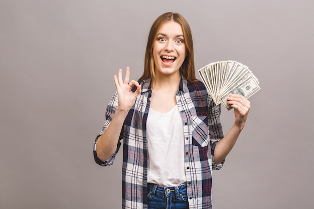 Porträt einer verspielten jungen Frau mit langen Haaren, die Bündel Geldbanknoten halten und Kamera betrachten. Ok Zeichen.