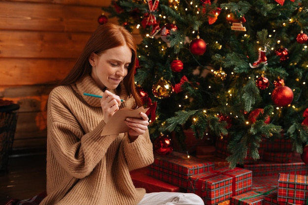 Porträt einer verträumten rothaarigen jungen Frau, die dem Weihnachtsmann eine Wunschliste für das neue Jahr schreibt