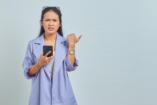 Porträt einer verwirrten jungen Asiatin, die Handy hält und mit dem Finger auf den Kopienraum zeigt, isoliert auf weißem Hintergrund
