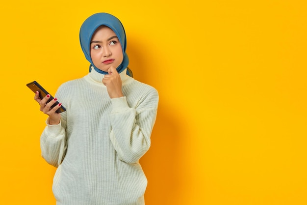 Porträt einer verwirrten jungen asiatischen Frau in weißem Pullover und Hijab, die mit der Hand am Kinn über Fragen nachdenkt, während sie das Mobiltelefon isoliert über gelbem Hintergrund hält Menschen islam religiöses Konzept