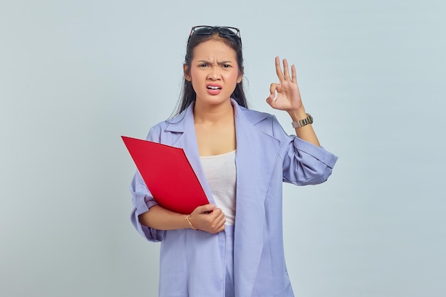 Porträt einer verwirrten jungen asiatischen Geschäftsfrau im Anzug, die einen Dokumentenordner hält und eine okay-Geste macht, stimmt mit einer netten Idee überein, die auf violettem Hintergrund isoliert ist