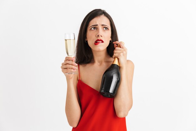 Porträt einer verwirrten jungen Frau im Kleid, das Glas und eine Flasche Champagner lokalisiert hält