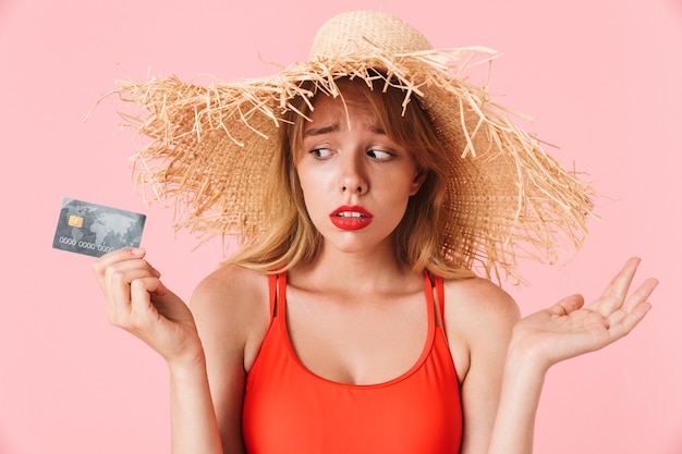 Porträt einer verwirrten jungen Frau mit langen lockigen Haaren mit Sommerstrohhut, die Kreditkarte isoliert über rosafarbener Wand hält