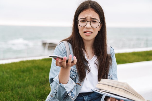 Porträt einer verwirrten unzufriedenen emotionalen jungen hübschen Studentin, die eine Brille trägt und im Freien ein Buch mit Handy liest.