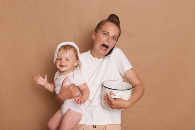 Porträt einer verzweifelten Frau, die ein weißes T-Shirt trägt, das ihre kleine Tochter in den Händen hält und Topf kocht, während das Kleinkind mit einem Telefon spricht und schreiend posiert, isoliert vor braunem Hintergrund