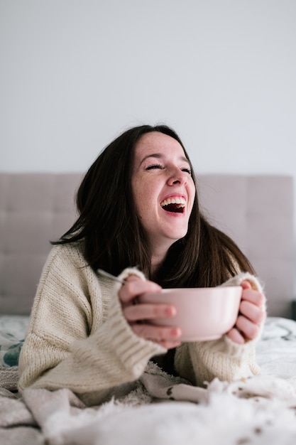 Porträt einer weißen Frau mit Sommersprossen und dunklem Haar, die laut auf dem Bett liegend lacht, während sie eine Schüssel hält