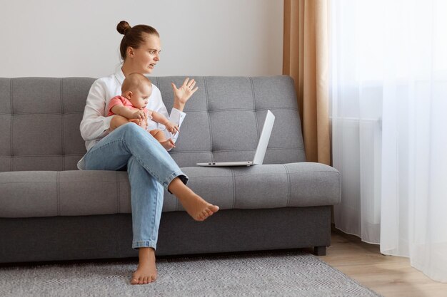 Porträt einer wütenden, aggressiven kaukasischen Frau in weißem T-Shirt, die mit ihrer kleinen Tochter auf dem Sofa sitzt und am Laptop arbeitet und Probleme mit der Arbeit hat, hat einen Fehler, der schreit und die Hand ausbreitet