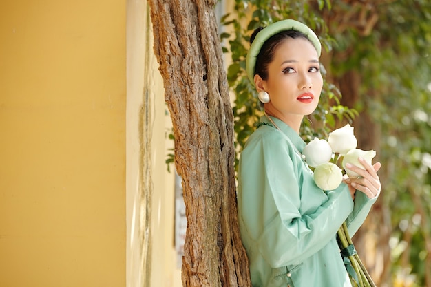 Porträt einer wunderschönen jungen vietnamesischen Frau in Ao Dai-Kleid, die weiße Lotusblumen hält und wegschaut