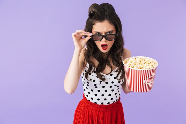 Porträt einer wunderschönen Pin-up-Frau in Retro-Polka-Dot-Kleid und 3D-Brille, die einen Eimer mit Popcorn hält, während sie einen Film einzeln über einer violetten Wand sieht