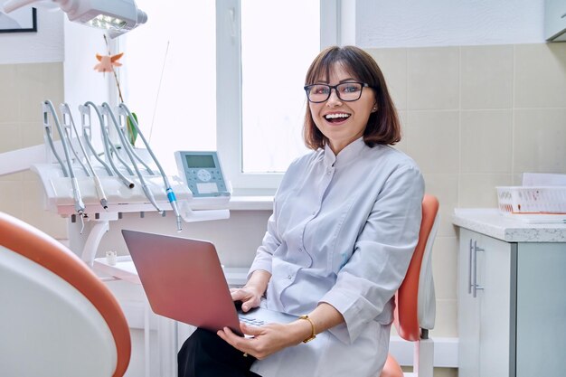 Porträt einer Zahnärztin, die mit einem Laptop in der Hand im Büro sitzt und in die Kamera blickt. Reife lächelnde Krankenschwester in der Zahnklinik. Zahnmedizin, Gesundheitsberuf, Stomatologie-Konzept