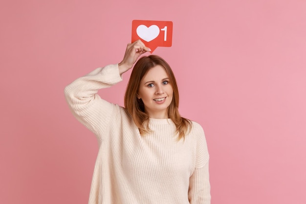 Porträt einer zufriedenen blonden Frau, die ein Social-Media-Herz wie das Symbol über dem Kopf hält und der Kamera lächelt, folgen und Inhalt tragen, der einen weißen Pullover trägt Innenstudioaufnahme isoliert auf rosa Hintergrund