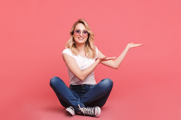 Porträt einer zufriedenen blonden Frau mit Vintage-Sonnenbrille lachend auf dem Boden sitzend mit gekreuzten Beinen isoliert über rosa Wand