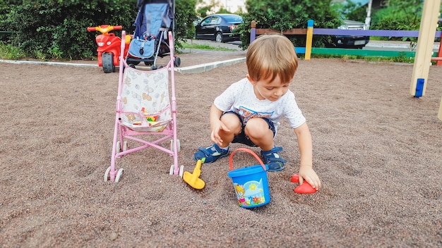 Porträt eines 3 Jahre alten Kleinkindjungen, der auf dem Spielplatz sitzt und Sand mit Plastikspaten und Eimer gräbt