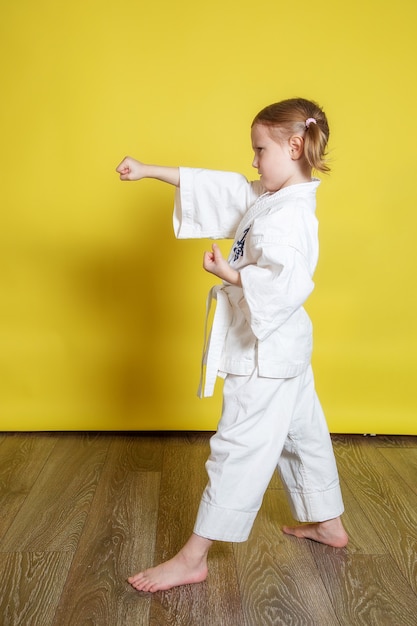 Porträt eines 5-jährigen Mädchens im Kimono, das zu Hause Karate gegen die gelbe Oberfläche praktiziert