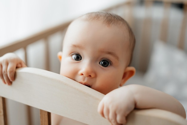Porträt eines 5 Monate alten Mädchens, das nach dem Schlafen in einer Krippe in einem hellen Kinderzimmer steht und in die Kamera schaut und lächelt