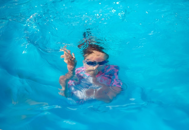 Porträt eines 8-jährigen Schwimmermädchens in einem hellen Badeanzug und einer blauen Brille in einem Freibad mit blauem Wasser unter Wasser während des Tauchens