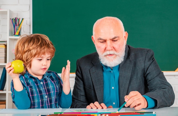 Porträt eines älteren Lehrers und eines kleinen Jungen, die im Klassenzimmer an einem Schreibtisch sitzen. Schüler und Lehrer im Klassezimmer.