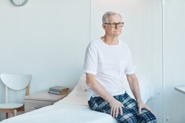 Porträt eines älteren Mannes, der auf einem Krankenhausbett im weißen Raum sitzt und nachdenklich wegschaut, Platz kopieren