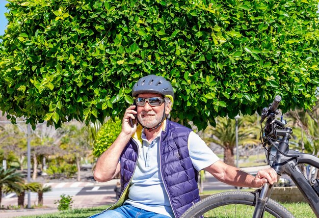 Porträt eines älteren Mannes mit Elektrofahrrad, der mit dem Handy im Freien im Park sitzt Lächelnder älterer Großvater, der einen Helm in der Nähe seines Fahrrads trägt und am Handy spricht