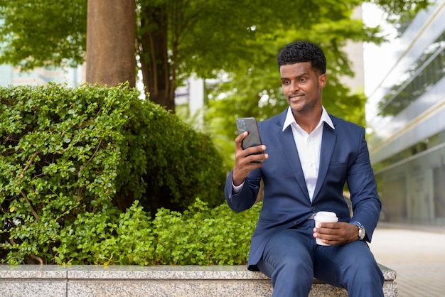 Porträt eines afrikanischen Geschäftsmannes, der Telefon benutzt und Kaffeetasse draußen in der Stadt hält, horizontaler Schuss