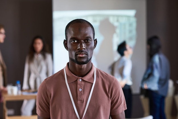 Porträt eines afrikanischen jungen Geschäftsmannes, der die Kamera anschaut, während er im Sitzungssaal mit anderen Leuten im Hintergrund steht