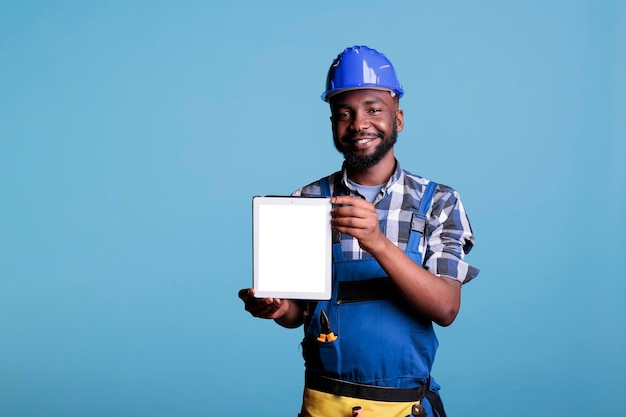 Porträt eines afroamerikanischen Baumeisters, der einen Tablet-Computer mit weißem Bildschirm hält, der im Studio mit blauem Hintergrund steht. Bauarbeiter, der Platz für Werbekopien empfiehlt.