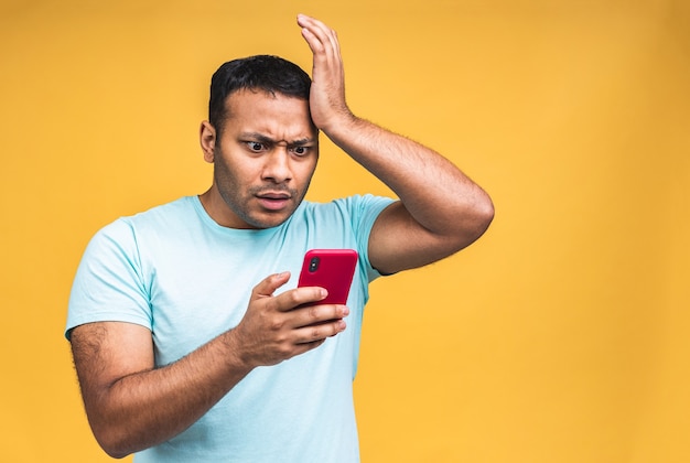 Porträt eines afroamerikanischen indischen schwarzen Mannes, schockiert, überrascht, weit geöffneter Mund, wütend auf das, was er auf seinem Handy sieht, einzeln auf gelbem Hintergrund. Negative menschliche Emotionen, Gesichtsausdrücke.