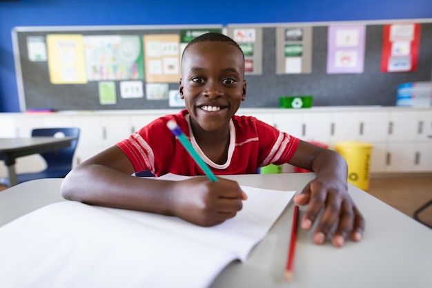 Porträt eines afroamerikanischen Jungen, der lächelt, während er in der Grundschulklasse auf seinem Schreibtisch sitzt