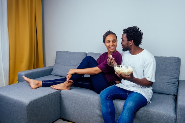 Porträt eines afroamerikanischen jungen Paares, das auf dem Sofa sitzt und einen Film sieht und Popcorn im Wohnzimmer isst.