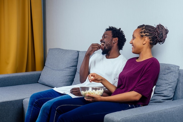 Porträt eines afroamerikanischen jungen Paares, das auf dem Sofa sitzt und einen Film sieht und Popcorn im Wohnzimmer isst.