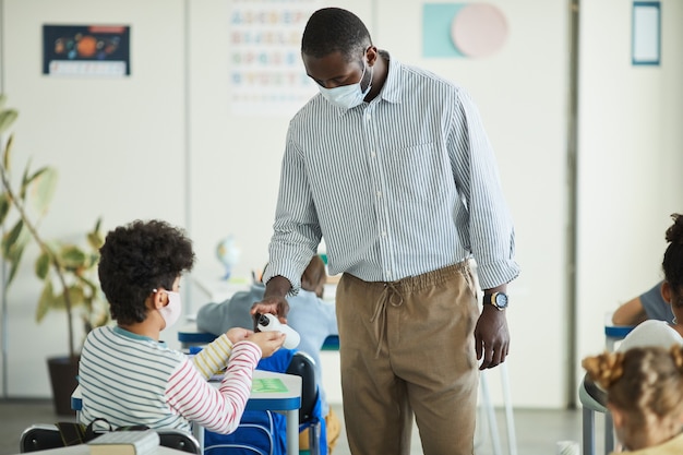 Porträt eines afroamerikanischen Lehrers, der die Hände von Kindern im Schulklassenzimmer desinfiziert, Kovid-Sicherheitsmaßnahmen, Kopierraum