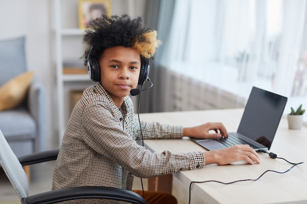 Porträt eines afroamerikanischen Teenagers, der ein Headset trägt und die Kamera anschaut, während er zu Hause einen Laptop verwendet, ein junges Gamer- oder Blogger-Konzept, Kopierraum