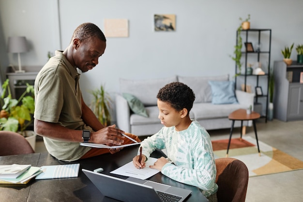 Porträt eines afroamerikanischen Vaters, der einem Teenager hilft, zu Hause im grünen Innenraum zu studieren