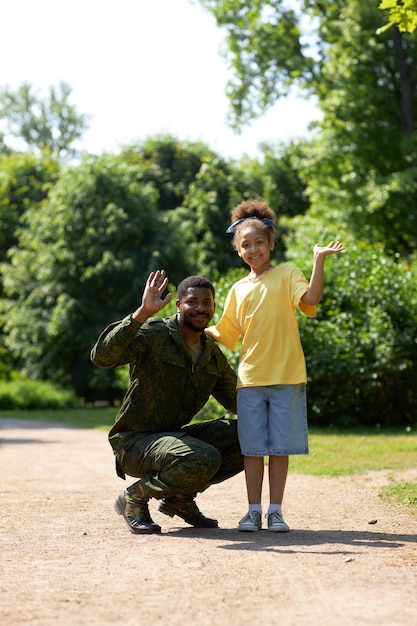 Porträt eines afroamerikanischen Vaters in Militäruniform, der zusammen mit seiner Tochter in die Kamera winkt