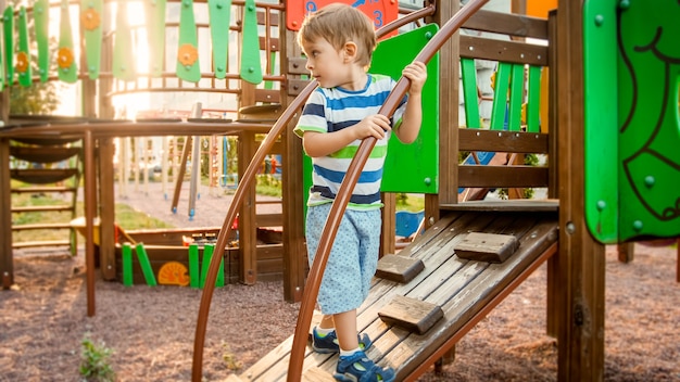 Porträt eines aktiven kleinen Jungen, der auf einer Holztreppe auf einem Kinderspielplatz im Park krabbelt und klettert