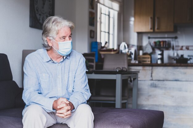 Porträt eines alten und reifen Mannes, der auf dem Sofa sitzt und eine Maske trägt, um Coronavirus in Innenräumen zu verhindern - Lockdown-Lifestyle zu Hause