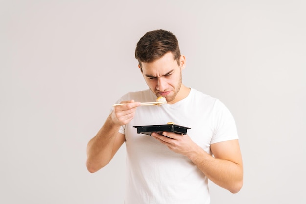 Porträt eines angewiderten jungen Mannes, der Sushi mit Essstäbchen hält und schnüffelt, mit einem Ausdruck von Abscheu im Gesicht auf weißem, isoliertem Hintergrund. Trauriger Kaukasier, der mit asiatischem Essen von schlechter Qualität unzufrieden ist.