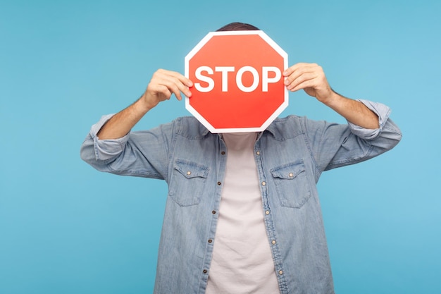 Foto porträt eines anonymen mannes im arbeiter-jeanshemd, das das gesicht mit dem verkehrszeichen stop als symbol für verbote, beschränkungen und verbotenen zugang bedeckt. studioaufnahme im innenbereich isoliert auf blauem hintergrund