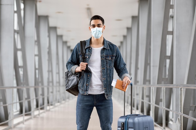 Porträt eines arabischen Mannes mit medizinischer Maske, der mit Gepäck am Flughafen steht