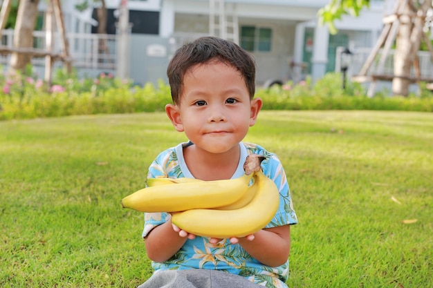 Porträt eines asiatischen kleinen Jungen im Alter von etwa 5 Jahren, der gelbe Bananen im Garten hält