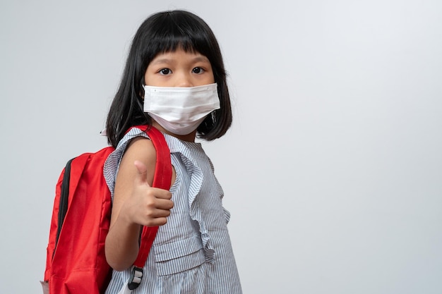 Porträt eines asiatischen Mädchenkindes mit schützender Gesichtsmaske und Schulrucksack und Daumen hoch bereit für das neue Schuljahr Konzept des Kindes, das zurück zur Schule geht und einen neuen normalen Lebensstil