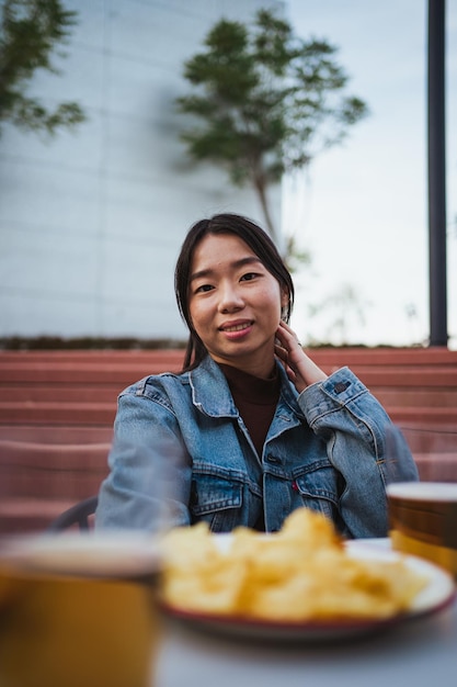 Porträt eines asiatischen Mädchens, das in die Kamera schaut, während sie ein paar Biere und Snacks auf einer Barterrasse trinkt