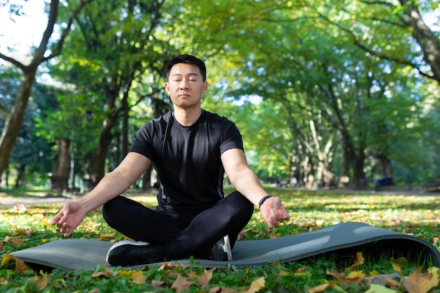 Porträt eines asiatischen Mannes im Herbstpark, der in Lotusposition auf einem Fitnessmattensportler sitzt und meditiert