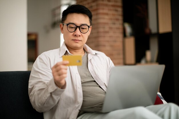 Porträt eines asiatischen reifen Mannes mit PC und Kreditkarte, der zu Hause auf der Couch sitzt, freier Speicherplatz