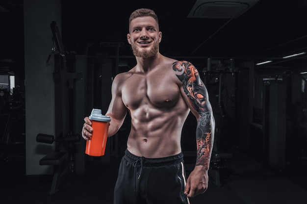 Porträt eines Athleten, der mit einem Shaker in der Turnhalle steht. Bodybuilding- und Fitnesskonzept. Gemischte Medien