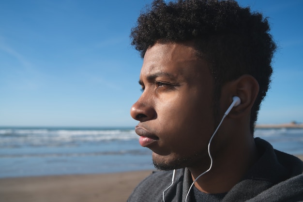 Porträt eines athletischen Mannes, der Musik mit Kopfhörern hört, während für Übung am Strand ruht. Sport und gesunder Lebensstil.