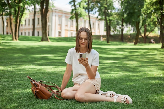 Porträt eines attraktiven, fröhlichen Mädchens in T-Shirt und kurzen Hosen, das auf Gras sitzt und mithilfe einer Geräte-App im Internet surft und seine Freizeit im Park im Freien verbringt