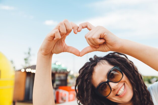 Porträt eines attraktiven, fröhlichen Mädchens mit gewellten Hairdreadlocks, das sich im Freien amüsiert und draußen ein Herzschild zeigt