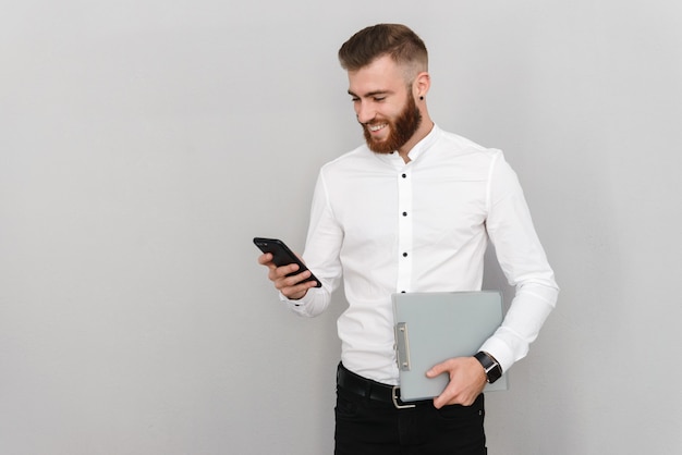 Porträt eines attraktiven, gutaussehenden jungen Geschäftsmannes, der über grauer Wand steht und SMS auf dem Handy schreibt, während er einen Laptop hält