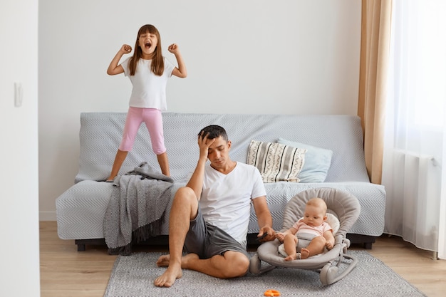 Porträt eines attraktiven jungen erwachsenen Mannes, der mit seinem kleinen Mädchen im Schaukelstuhl auf dem Boden in der Nähe des Sofas sitzt, müde von der lauten älteren Tochter ist, Kopfschmerzen hat, weißes T-Shirt und kurze Jeans trägt.